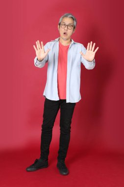 Kırmızı arka planda duran mavi tişörtlü, uzun saçlı kıdemli Asyalı adam..