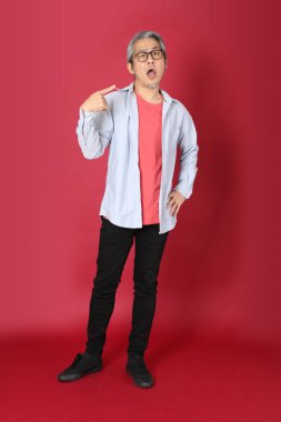 Kırmızı arka planda duran mavi tişörtlü, uzun saçlı kıdemli Asyalı adam..