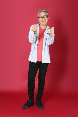 Kırmızı arka planda duran mavi tişörtlü, uzun saçlı kıdemli Asyalı adam..