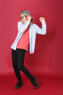 Kırmızı arka planda duran mavi tişörtlü, uzun saçlı kıdemli Asyalı adam..