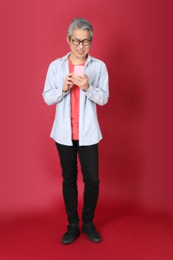 Kırmızı arka planda duran mavi tişörtlü, uzun saçlı kıdemli Asyalı adam..