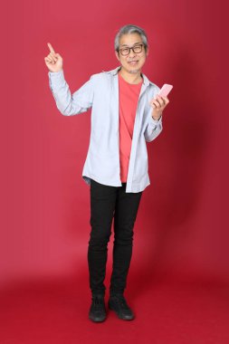 Kırmızı arka planda duran mavi tişörtlü, uzun saçlı kıdemli Asyalı adam..