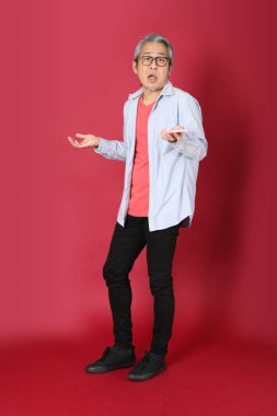 Kırmızı arka planda duran mavi tişörtlü, uzun saçlı kıdemli Asyalı adam..