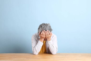 The senior Asian man with grey shirt sitting at the working desk with the blue background.