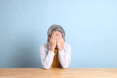 The senior Asian man with grey shirt sitting at the working desk with the blue background.