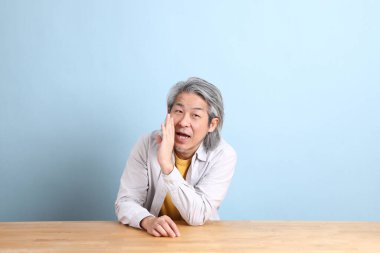 The senior Asian man with grey shirt sitting at the working desk with the blue background.
