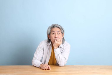 The senior Asian man with grey shirt sitting at the working desk with the blue background.