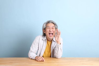 The senior Asian man with grey shirt sitting at the working desk with the blue background.