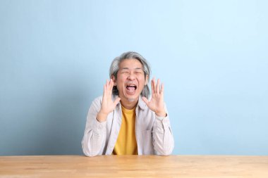 The senior Asian man with grey shirt sitting at the working desk with the blue background.