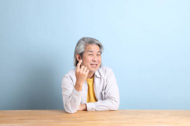 The senior Asian man with grey shirt sitting at the working desk with the blue background.