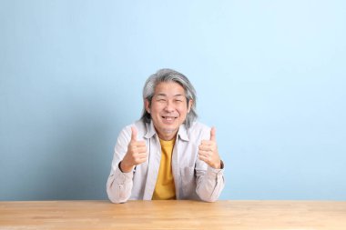 The senior Asian man with grey shirt sitting at the working desk with the blue background.