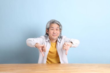 The senior Asian man with grey shirt sitting at the working desk with the blue background.