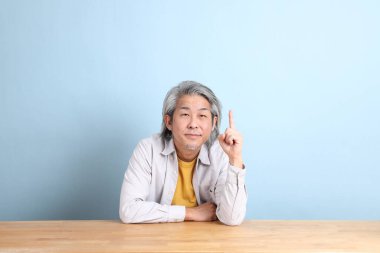 The senior Asian man with grey shirt sitting at the working desk with the blue background.