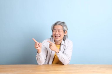 The senior Asian man with grey shirt sitting at the working desk with the blue background.