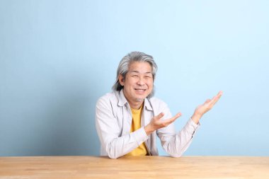 The senior Asian man with grey shirt sitting at the working desk with the blue background.
