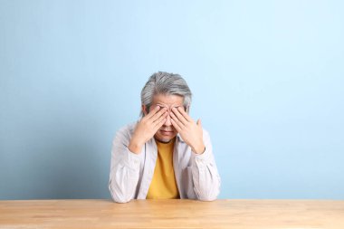 The senior Asian man with grey shirt sitting at the working desk with the blue background.