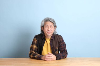 The senior Asian man wearing the yellow plaid shirt sitting at the working desk with blue background.