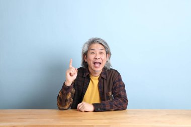The senior Asian man wearing the yellow plaid shirt sitting at the working desk with blue background.