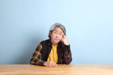 The senior Asian man wearing the yellow plaid shirt sitting at the working desk with blue background.