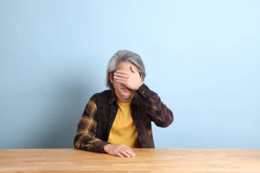 The senior Asian man wearing the yellow plaid shirt sitting at the working desk with blue background.