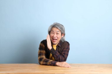 The senior Asian man wearing the yellow plaid shirt sitting at the working desk with blue background.