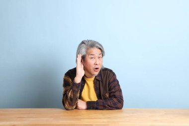 The senior Asian man wearing the yellow plaid shirt sitting at the working desk with blue background.