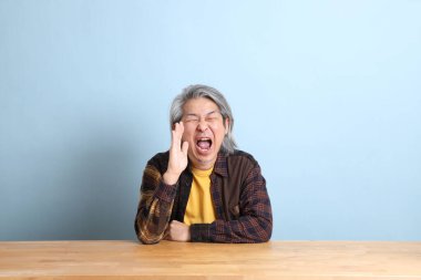 The senior Asian man wearing the yellow plaid shirt sitting at the working desk with blue background.