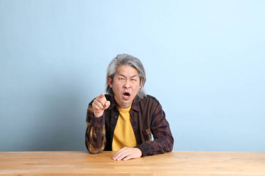 The senior Asian man wearing the yellow plaid shirt sitting at the working desk with blue background.
