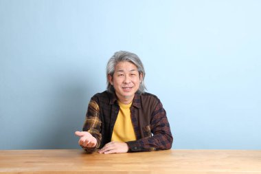 The senior Asian man wearing the yellow plaid shirt sitting at the working desk with blue background.