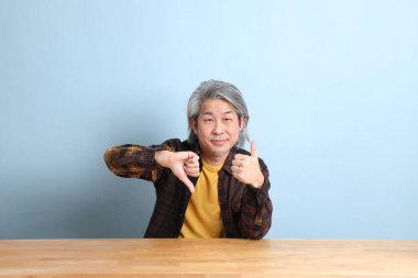 The senior Asian man wearing the yellow plaid shirt sitting at the working desk with blue background.