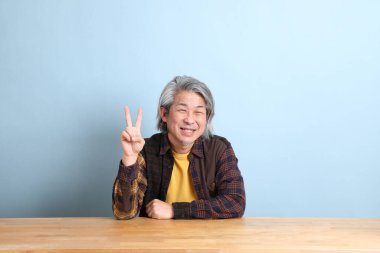 The senior Asian man wearing the yellow plaid shirt sitting at the working desk with blue background.