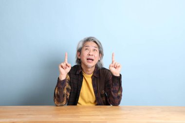 The senior Asian man wearing the yellow plaid shirt sitting at the working desk with blue background.