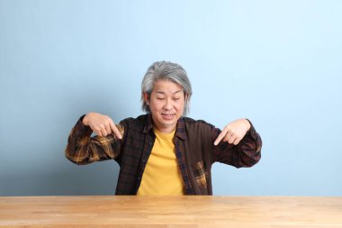 The senior Asian man wearing the yellow plaid shirt sitting at the working desk with blue background.