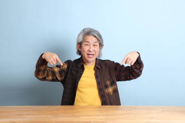 The senior Asian man wearing the yellow plaid shirt sitting at the working desk with blue background.