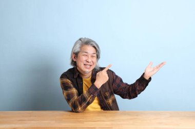 The senior Asian man wearing the yellow plaid shirt sitting at the working desk with blue background.