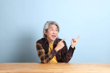 The senior Asian man wearing the yellow plaid shirt sitting at the working desk with blue background.