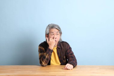 The senior Asian man wearing the yellow plaid shirt sitting at the working desk with blue background.