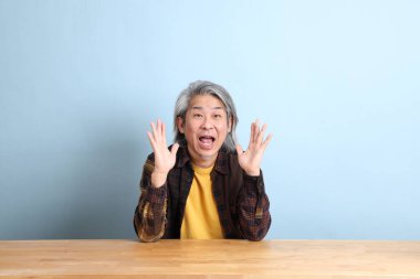 The senior Asian man wearing the yellow plaid shirt sitting at the working desk with blue background.
