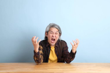 The senior Asian man wearing the yellow plaid shirt sitting at the working desk with blue background.