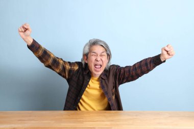 The senior Asian man wearing the yellow plaid shirt sitting at the working desk with blue background.