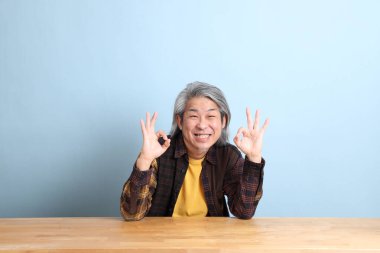 The senior Asian man wearing the yellow plaid shirt sitting at the working desk with blue background.