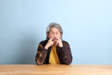 The senior Asian man wearing the yellow plaid shirt sitting at the working desk with blue background.