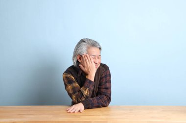 The senior Asian man wearing the yellow plaid shirt sitting at the working desk with blue background.