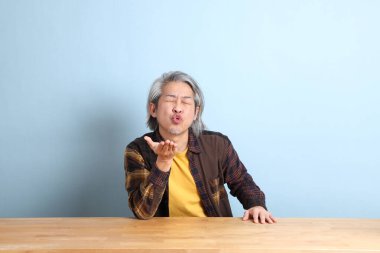 The senior Asian man wearing the yellow plaid shirt sitting at the working desk with blue background.