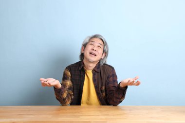 The senior Asian man wearing the yellow plaid shirt sitting at the working desk with blue background.