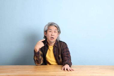 The senior Asian man wearing the yellow plaid shirt sitting at the working desk with blue background.