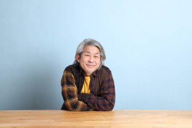 The senior Asian man wearing the yellow plaid shirt sitting at the working desk with blue background.