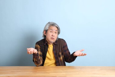 The senior Asian man wearing the yellow plaid shirt sitting at the working desk with blue background.