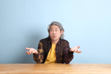 The senior Asian man wearing the yellow plaid shirt sitting at the working desk with blue background.