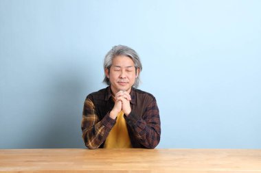 The senior Asian man wearing the yellow plaid shirt sitting at the working desk with blue background.